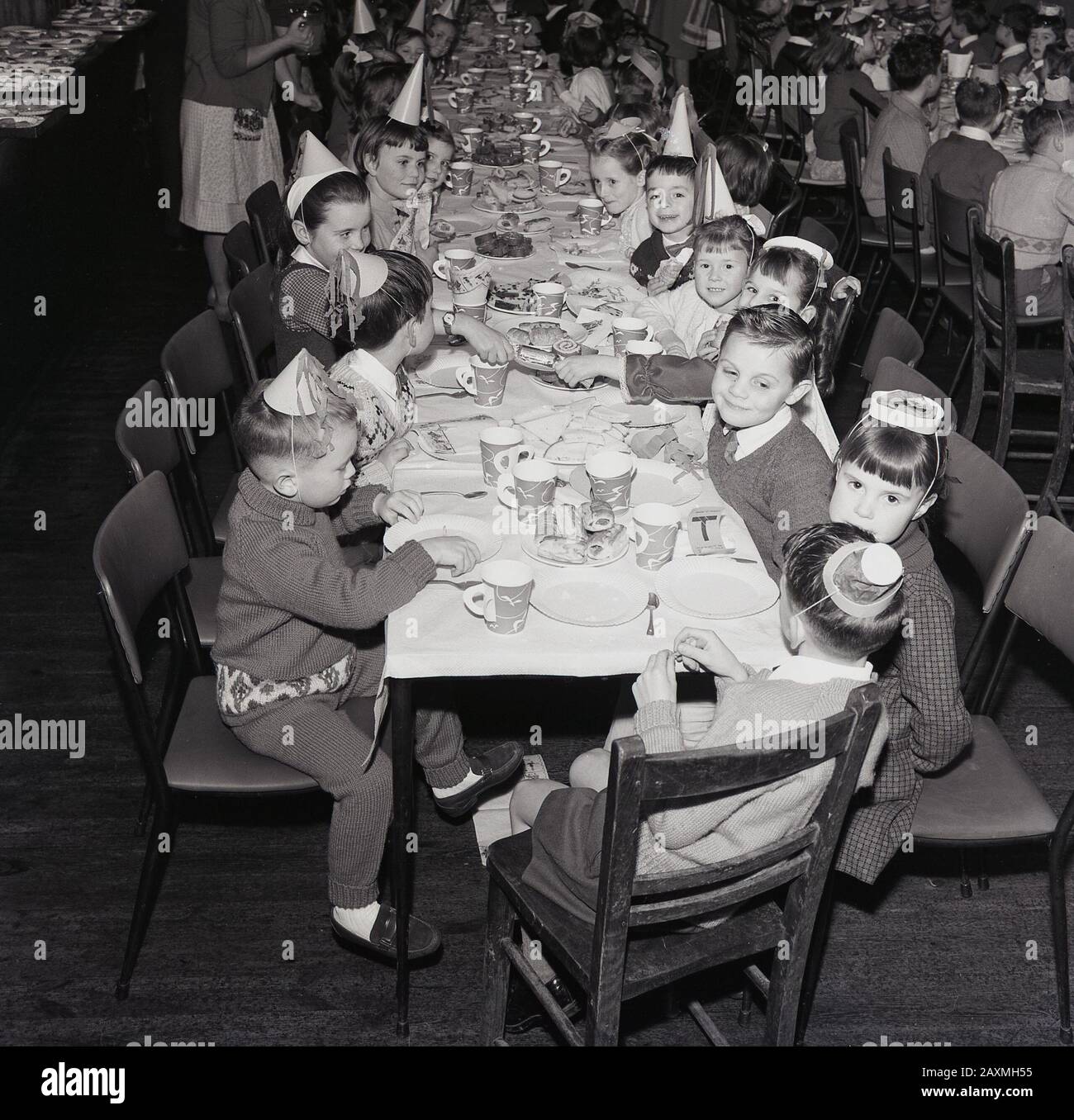 1960er Jahre, historisch, in einem Speisesaal, kleine Schulkinder in Partyhüten, die gemeinsam bei einem weihnachtsessen Spaß haben, England, Großbritannien Stockfoto