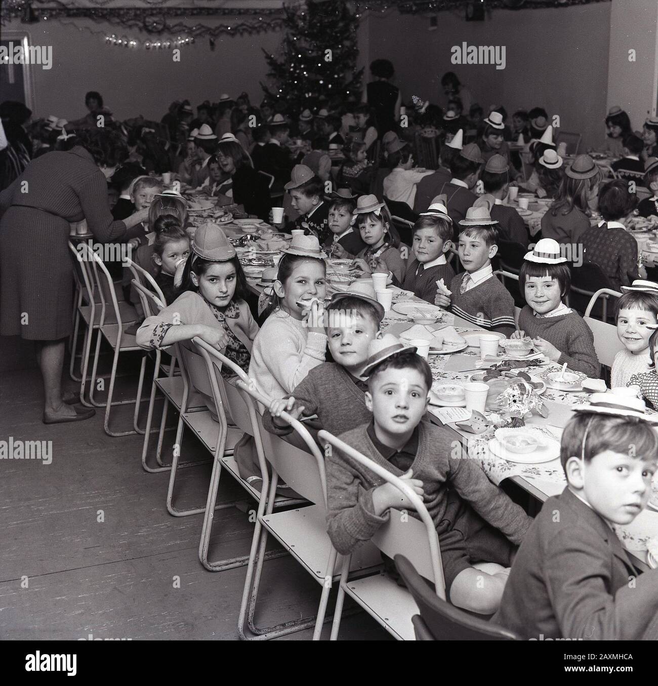 1960er Jahre, historisch, in einem Speisesaal, kleine Schulkinder in Partyhüten, die gemeinsam bei einem weihnachtsessen Spaß haben, England, Großbritannien. Stockfoto
