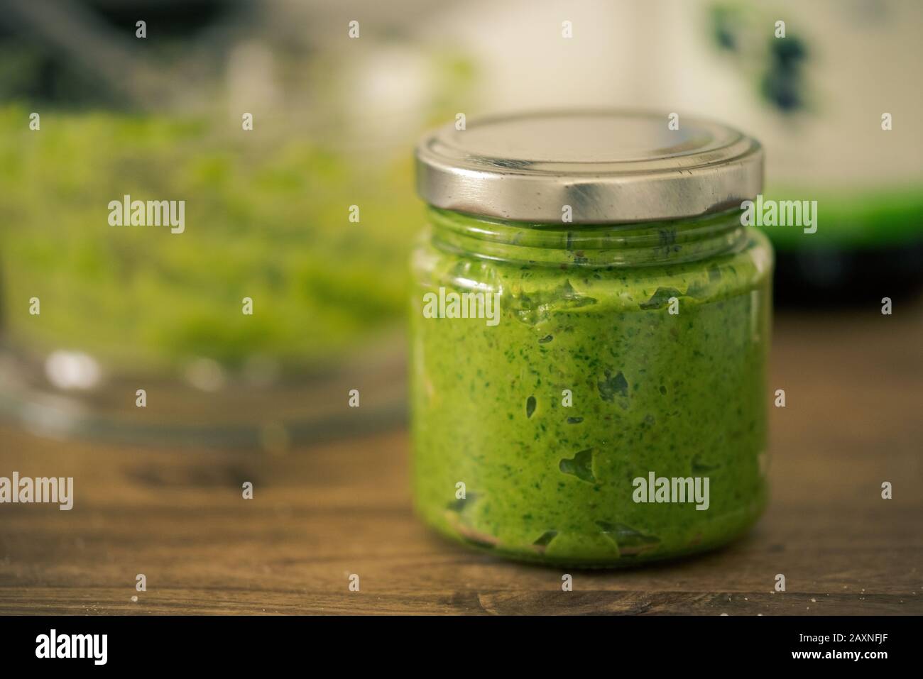 Grüner Pesto in wiederverwendbarem Schraubglas, das auf dunklem Holztisch mit unscharfem Hintergrund steht Stockfoto