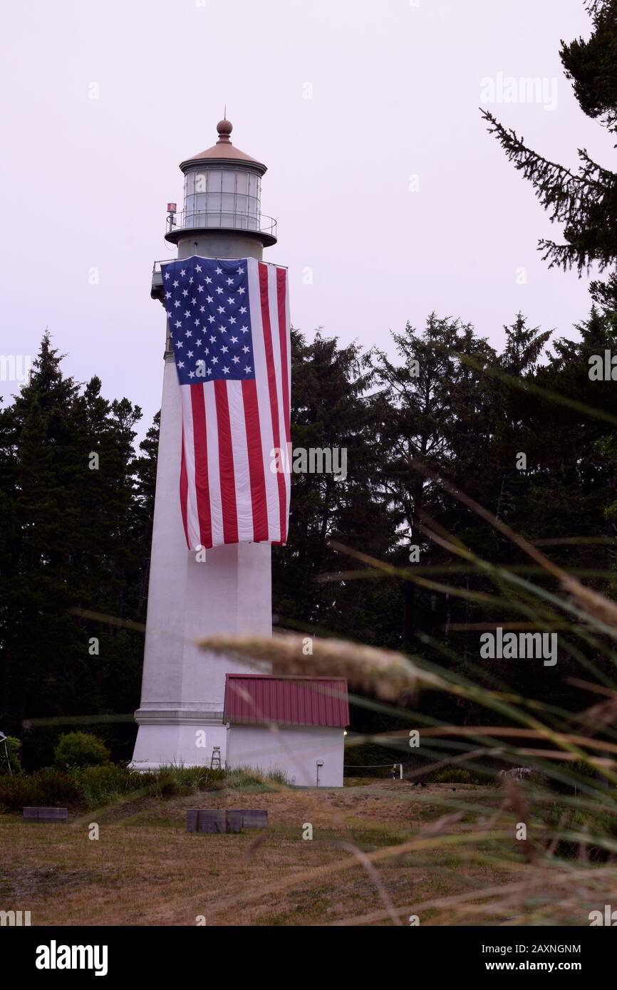 Wunderschöner Leuchtturm Mit Amerikanischer Flagge Umgeben Von Evergreen Forest Gegen Bewölkten Morgen-Himmel Stockfoto