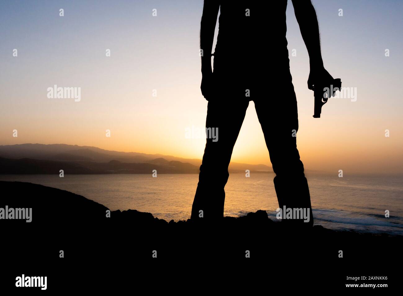 Silhouette des Mannes, der bei Sonnenuntergang auf Klippen mit Blick auf das Meer Pistole hält Stockfoto