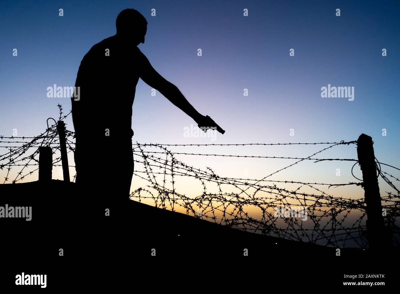 Silhouette des Mannes, der die Waffe in der Nähe des Stacheldrahtzauns zeigt. Stockfoto
