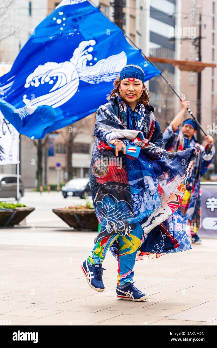 Junge Japanerin Yosakoi-Tänzerin, tanzt mit Naruko, Klappern, draußen auf dem Kyusyu Gassai Festival, Kumamoto. Mann winkt Banner im Hintergrund. Stockfoto