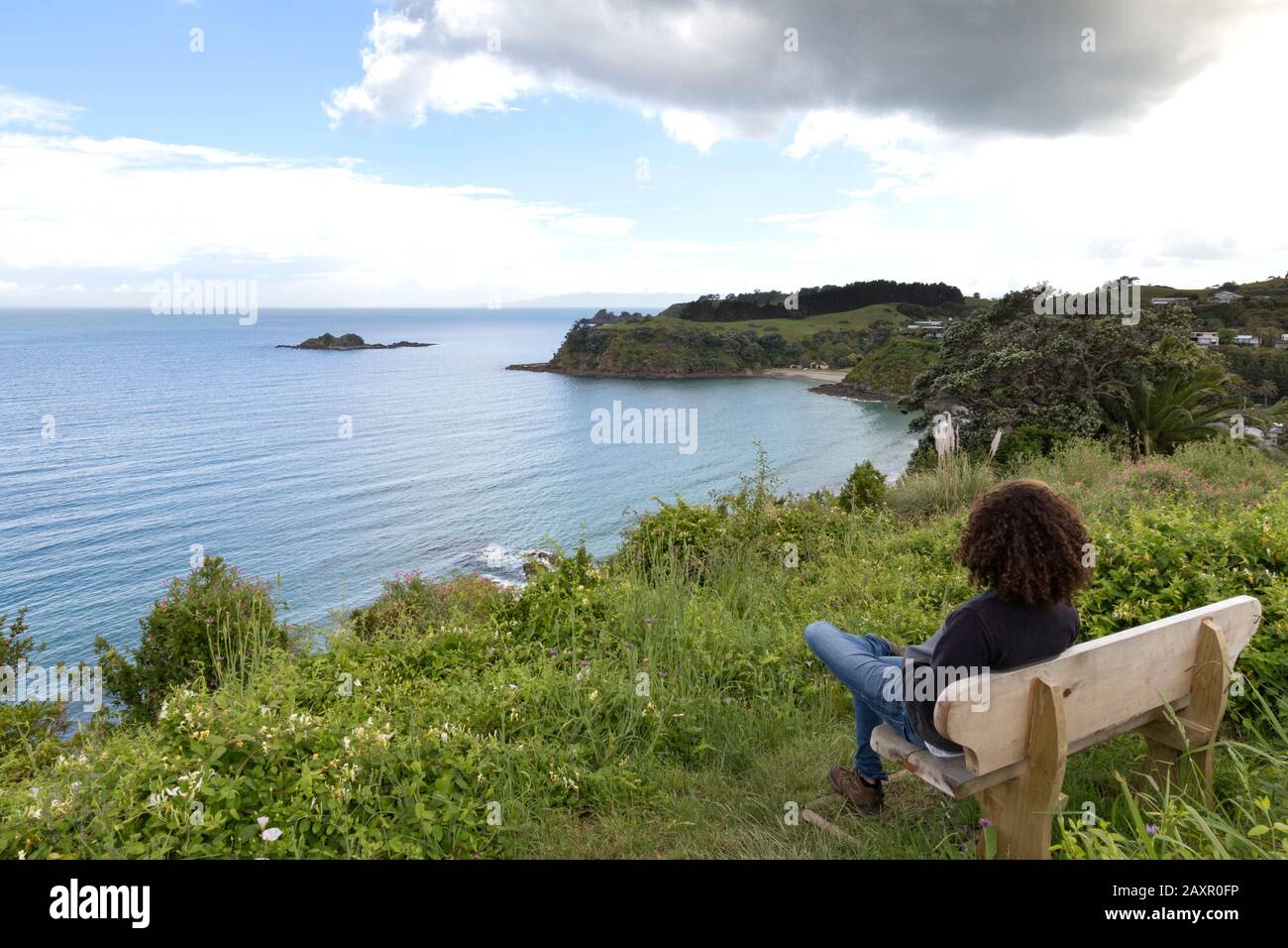 Mann mit lockigen Haaren, der auf das Meer blickt, sitzt auf einer Bank, ist Waiheke. Stockfoto