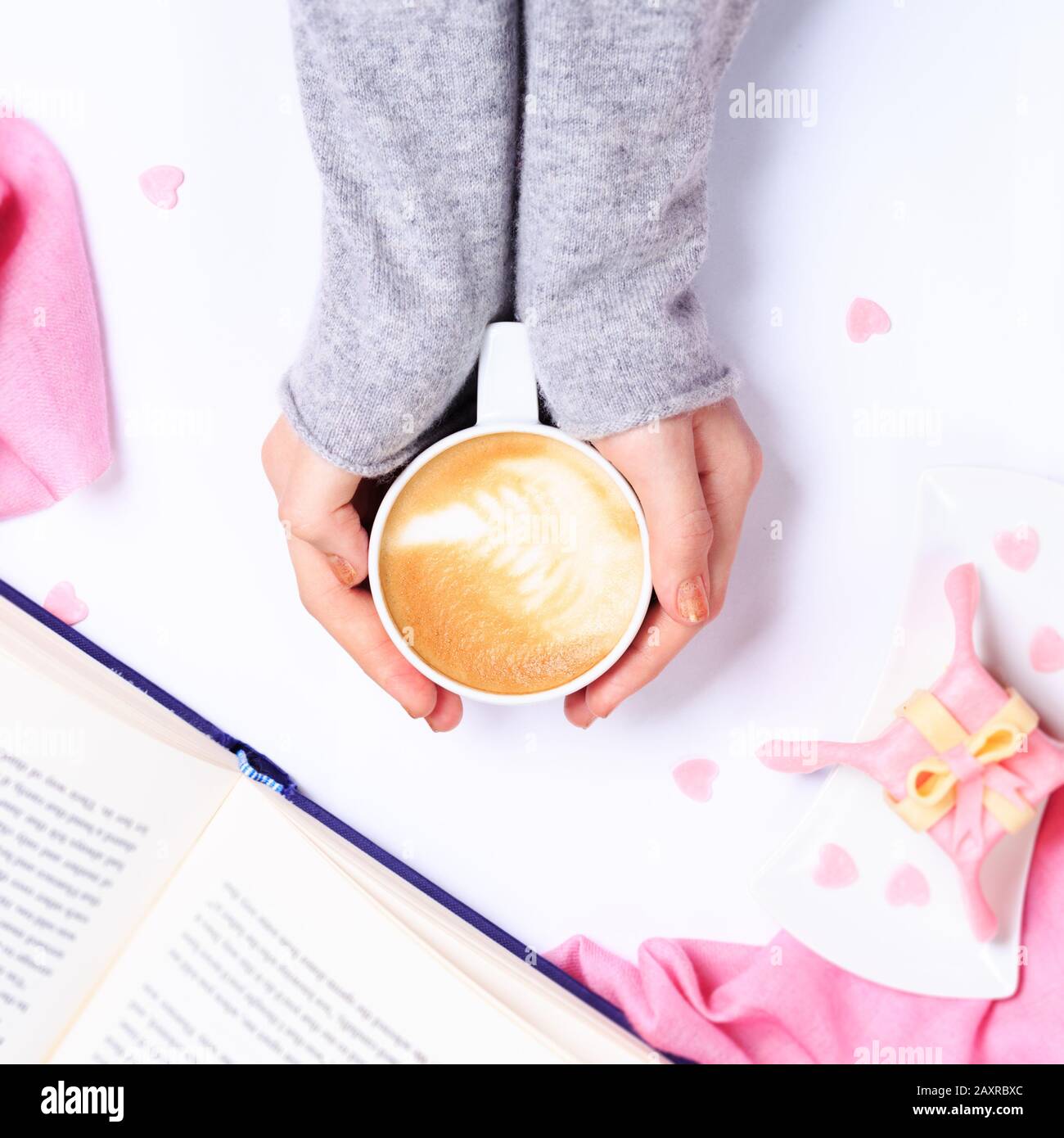 Weibliche Hände in grau gestricktem Haltebecher aus Cappuccino. Geschenk gleich Dessert mit herzförmigen Besprengungen. Geöffnetes Buch. Draufsicht. Valentines Konzept. Rosa Textil. Platz Erreichte Stockfoto