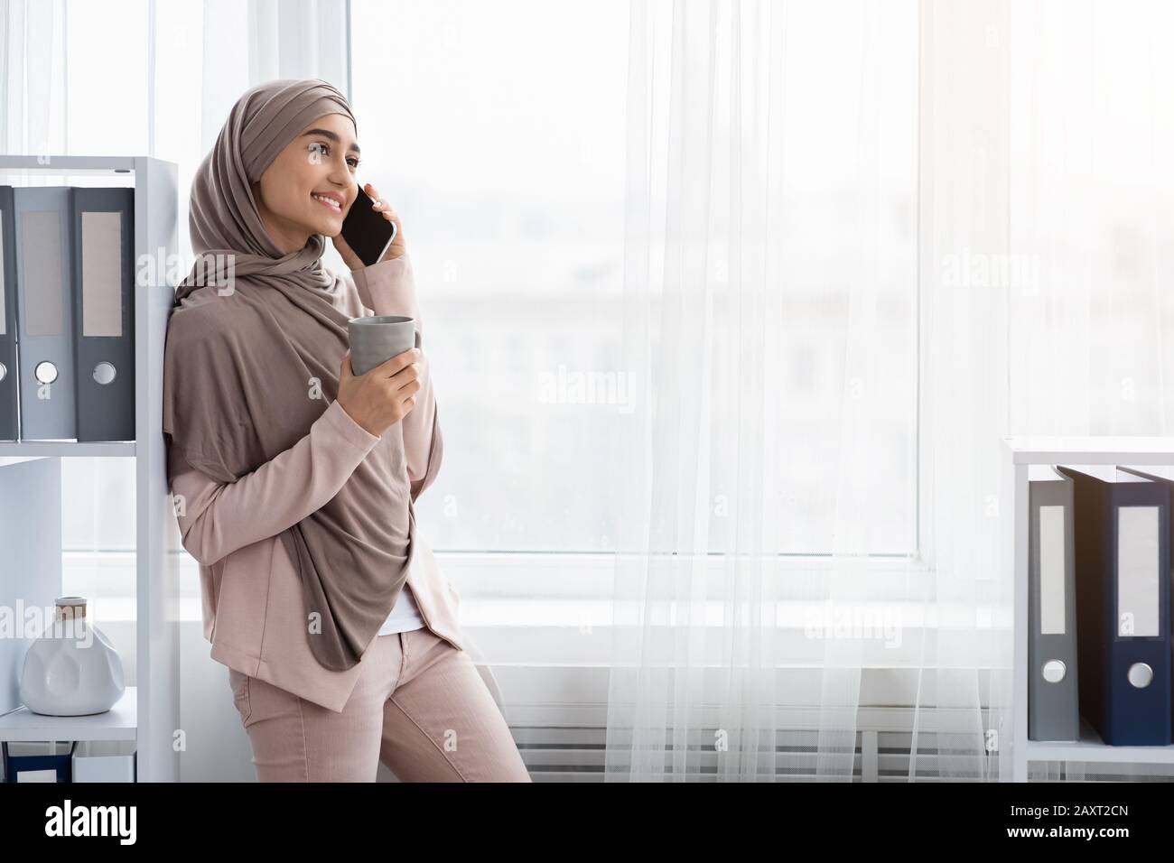 Junge muslimische Geschäftsfrau, die sich mit Smartphone und Kaffee im Büro entspannt Stockfoto