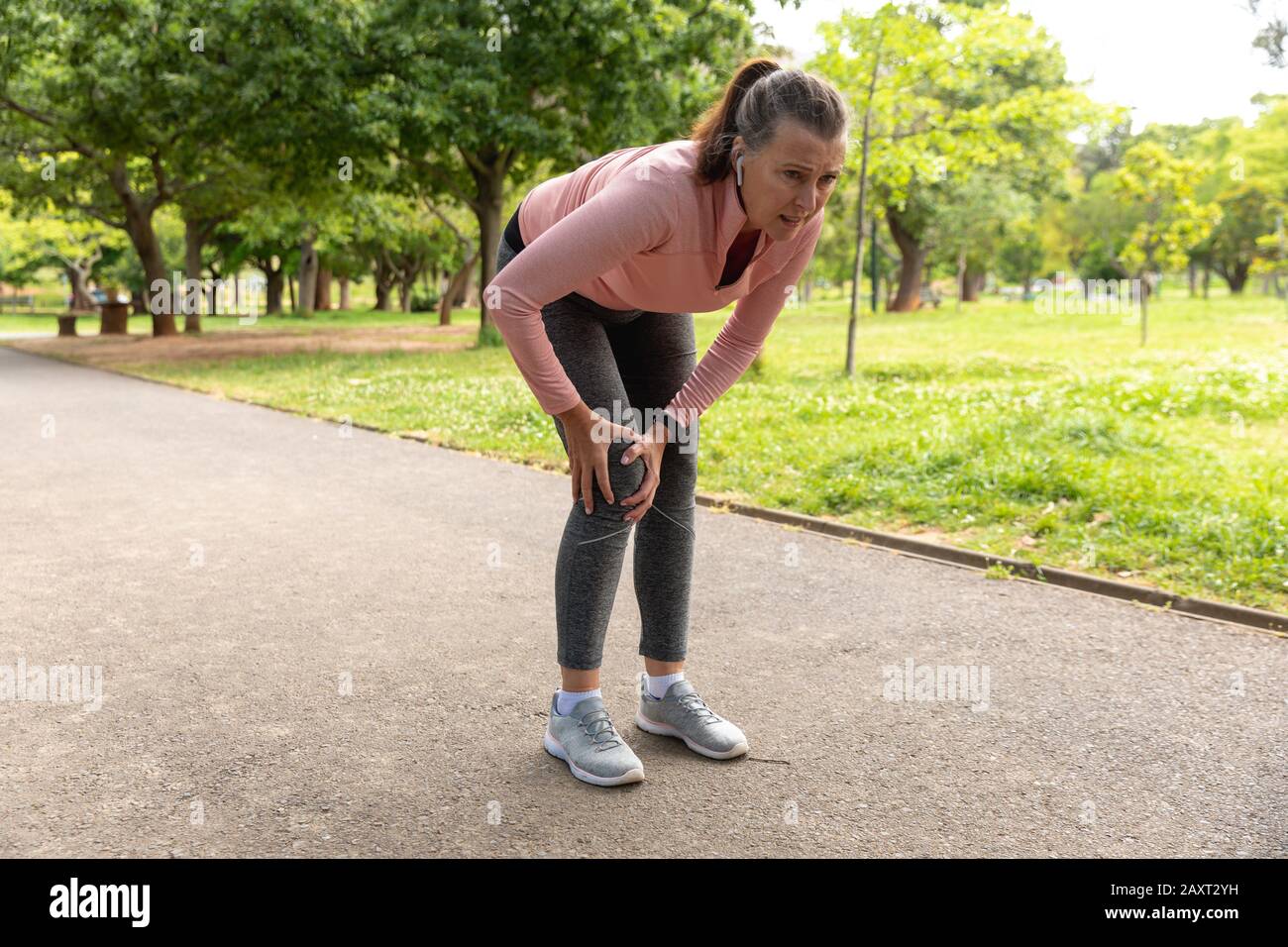 Seitenansicht einer Frau im mittleren Alter, die in einem Park ausarbeitet, ihr verletztes Knie hält und sich bei Schmerzen nach unten beugt Stockfoto