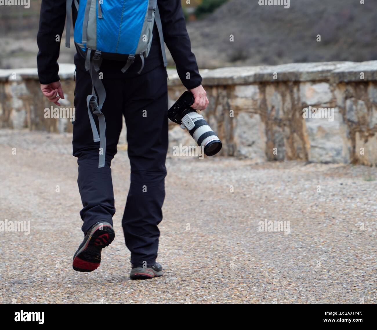 Mann mit blauem Beutel, der eine Kamera mit einem großen Zoomobjektiv im Freien inmitten eines Pfades hält, der von einer Steinmauer in Maderuelo in umgeben ist Stockfoto