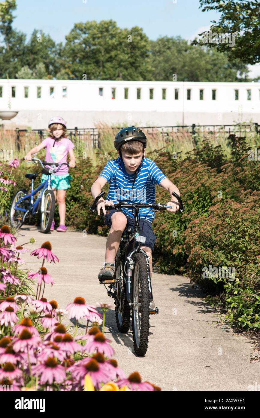 Ein elfjähriger Junge fährt mit dem Fahrrad im Beuter Riverwalk Park in Mishawaka, Indiana, USA, während seine neunjährige Schwester im Hintergrund beobachtet. Stockfoto