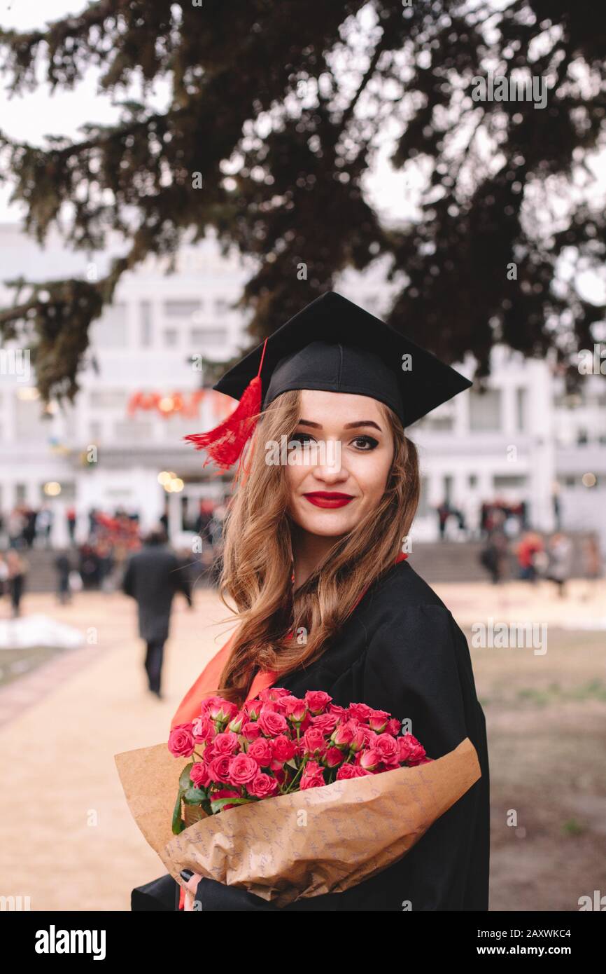 Fröhliche junge Studentin im Graduiertenkolleg, die Blumenstrauß hält, während sie bei der Abschlussfeier auf dem Campus steht Stockfoto