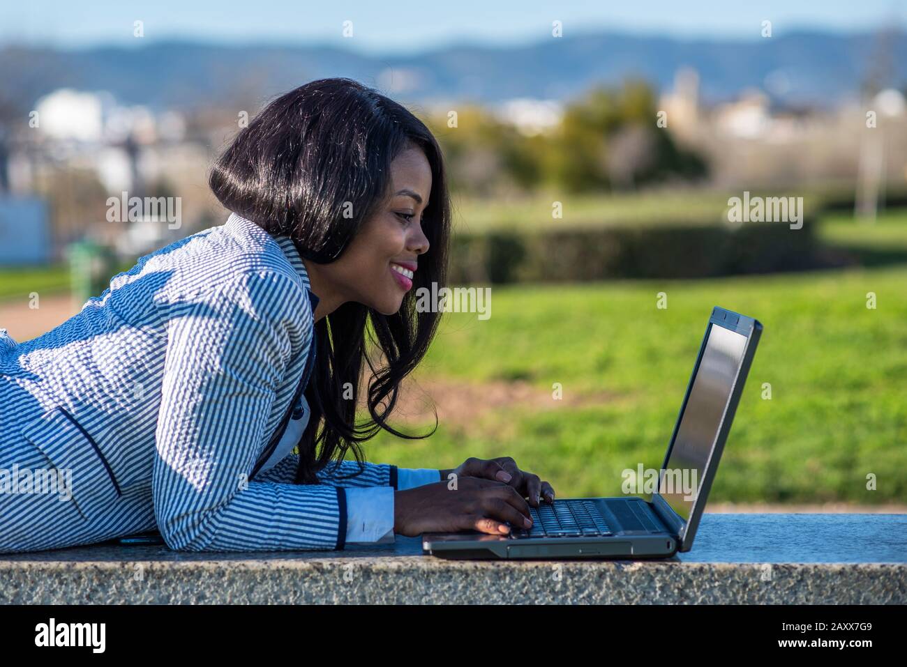 Lächelnde afro-amerikanische Frau, die an einem sonnigen Tag mit einem Laptop in einem Außenpark auf einer Bank liegt. Stockfoto