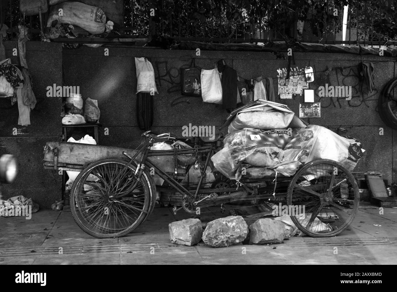 Die Wohnung einer in Neu-Delhi lebenden Straßenperson mit Fahrrad, Plastikblech und verschiedenen persönlichen Besitztümern, im Schatten in Schwarz-Weiß Stockfoto