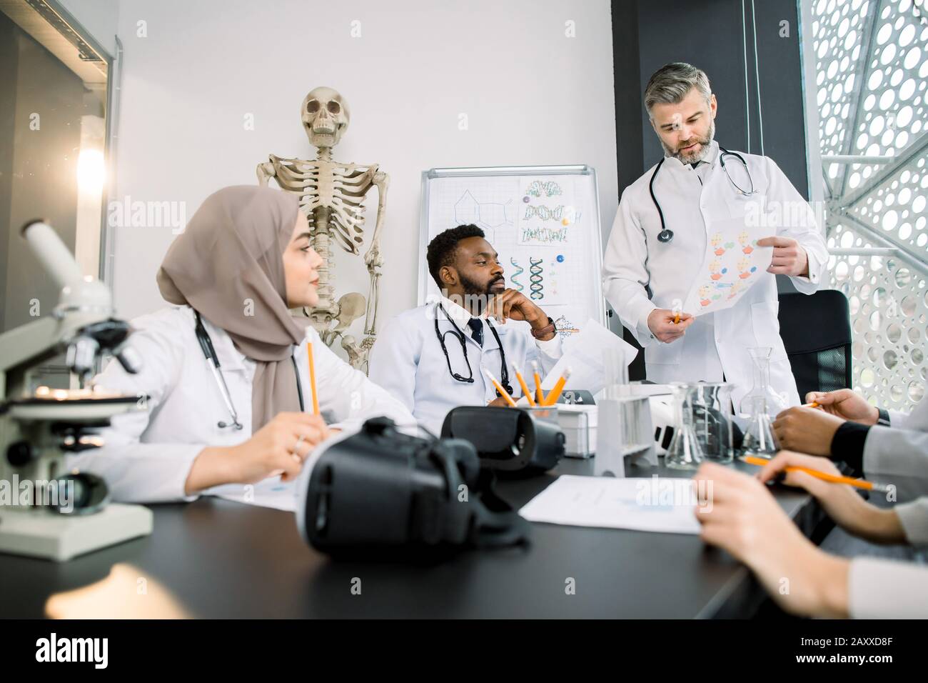 Eine Gruppe von multirassischen Medizinstudenten, die zusammen sitzen und studieren und sich während der Unterrichtsstunde mit ihrem erfahrenen männlichen Professor anlisten Stockfoto