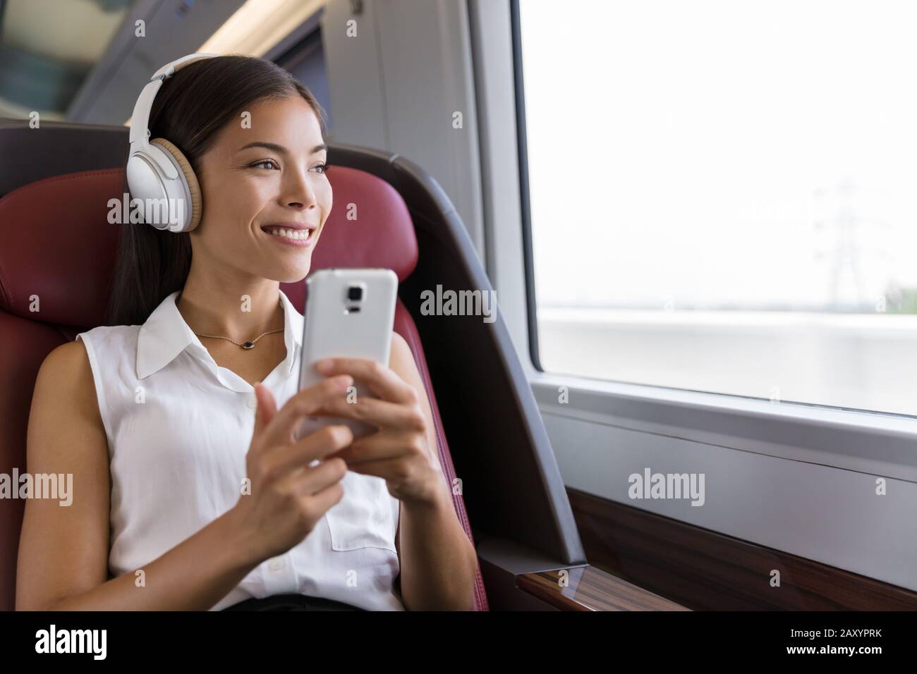 Frau im Zug, die Musik auf dem Smartphone hört. Lebensstil der Menschen. Junge Geschäftsfrau in der Stadt, die Telefon-App verwendet, drahtlose Kopfhörer, um Hörbücher zu hören. Asiatische Mädchen genießen Reisen in der Business Class. Stockfoto