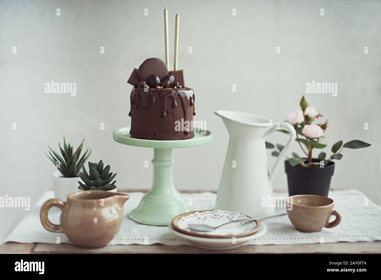 Schokoladenkuchen mit goldenen Kerzen auf einem Kakestand neben saftigen Pflanzen und Geschirr Stockfoto