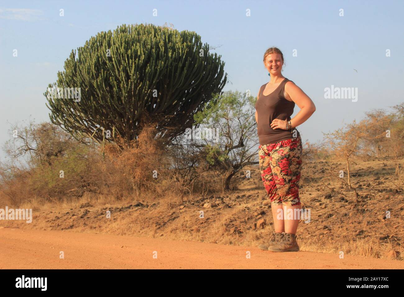 Weiße junge vollfette europäische Frau, die in der afrikanischen Savanne in Uganda vor dem Hintergrund eines riesigen Kakteens steht. Stockfoto