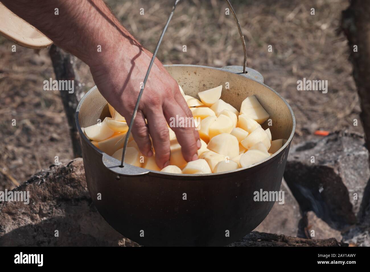 Die männliche Hand fügt in einem Kessel geschnittene Kartoffel hinzu. Zubereitung einer Suppe im offenen Feuer, Campingmahlzeit Stockfoto