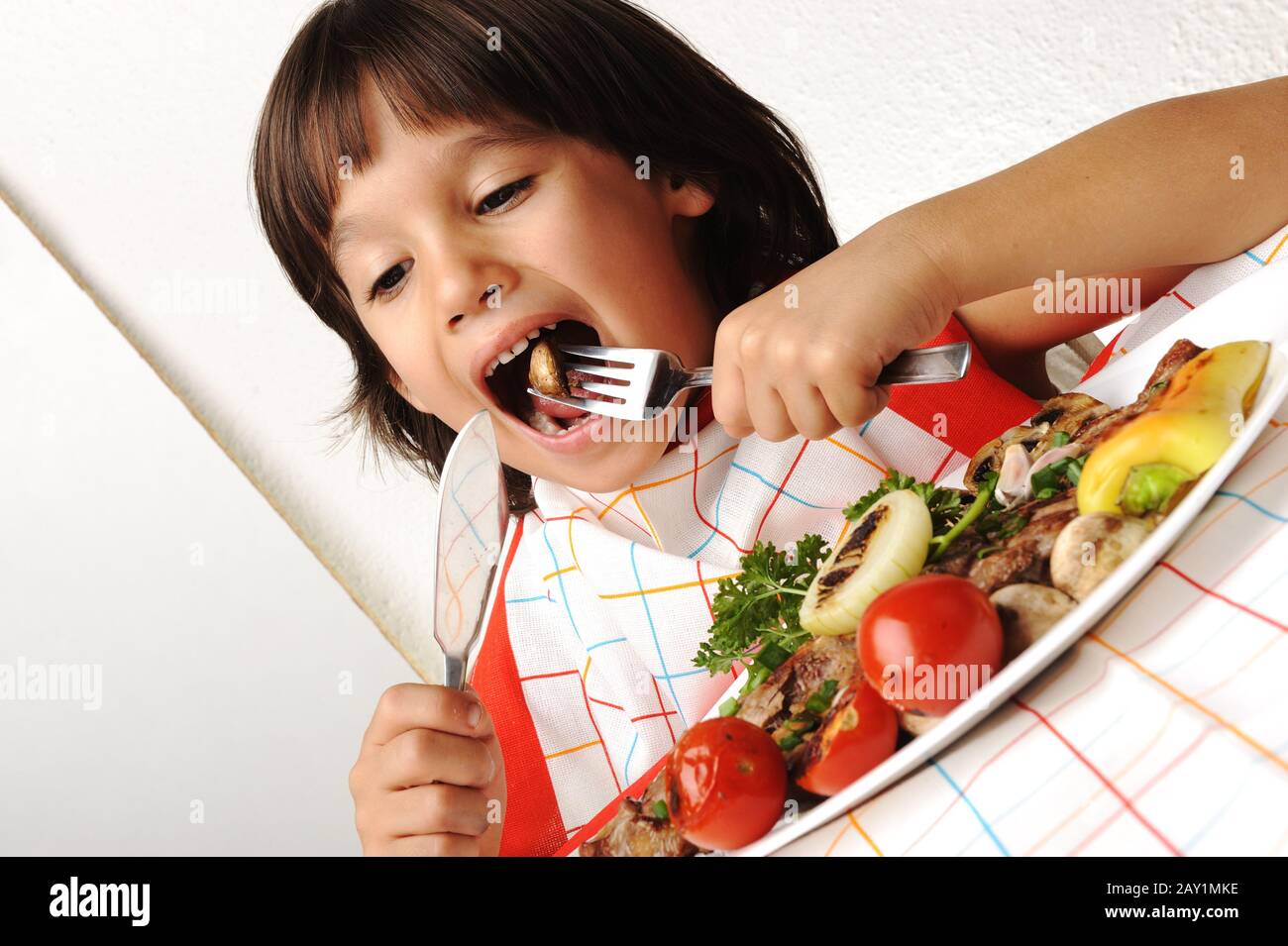 Süßer positiver Junge mit Gabel und Messer, der auf dem Mittagstisch isst Stockfoto