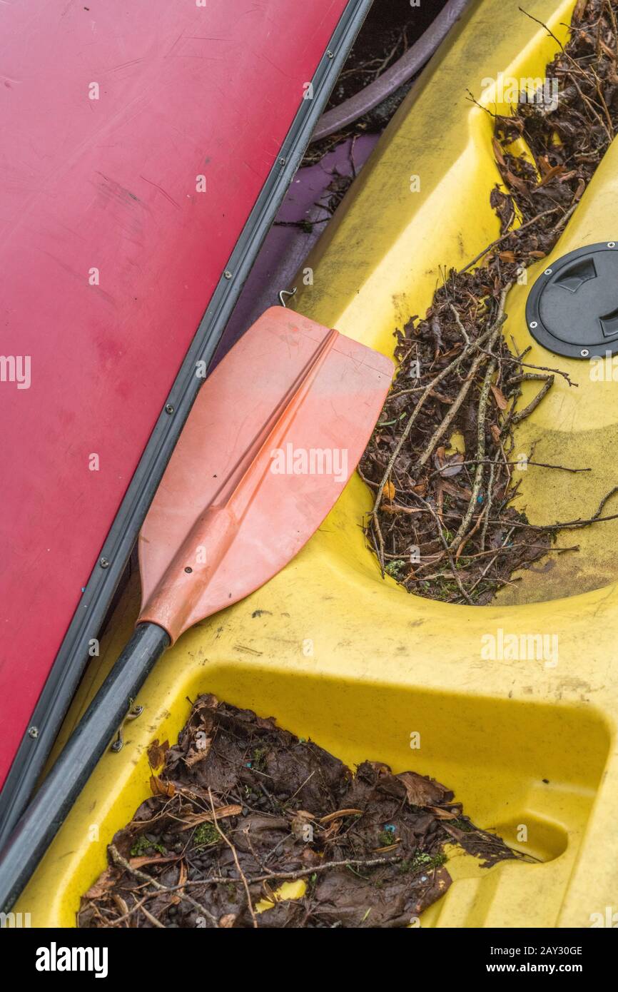 Zwei gebeutelte Kajaks aus Fiberglas am Flussufer, zusammen mit einem Kajaktaddel. Metapher für außerhalb der Saison, saisonale Aktivitäten, Kajakfahren. Stockfoto