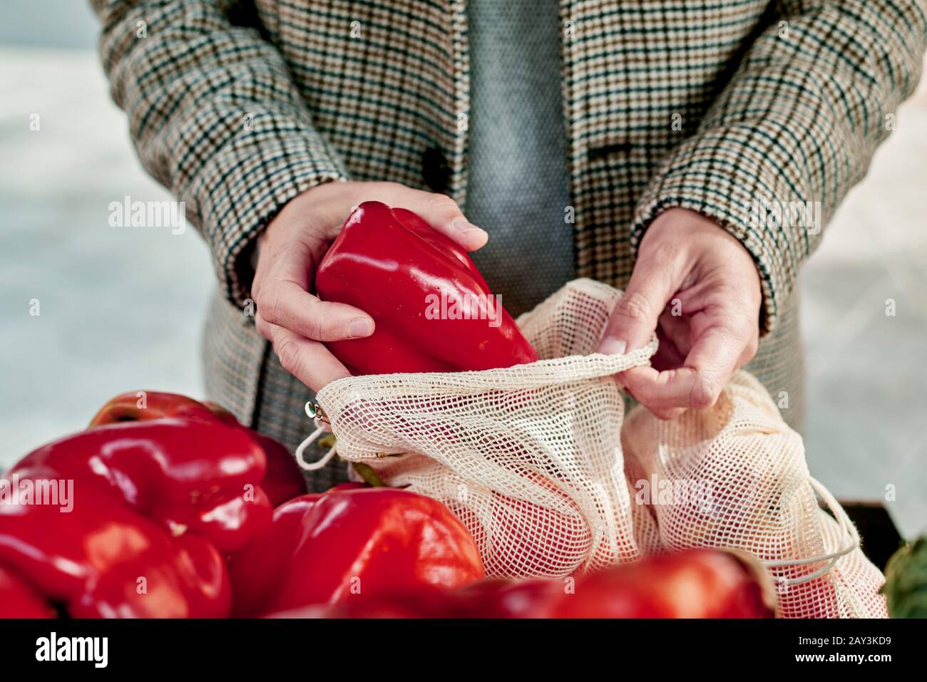 Nahaufnahme eines Mannes, der in einem Greengrocker einkauft und einige rote Paprika in einen wiederverwendbaren Textil-Netzbeutel stellt, um die Kunststoffverschmutzung zu verringern Stockfoto