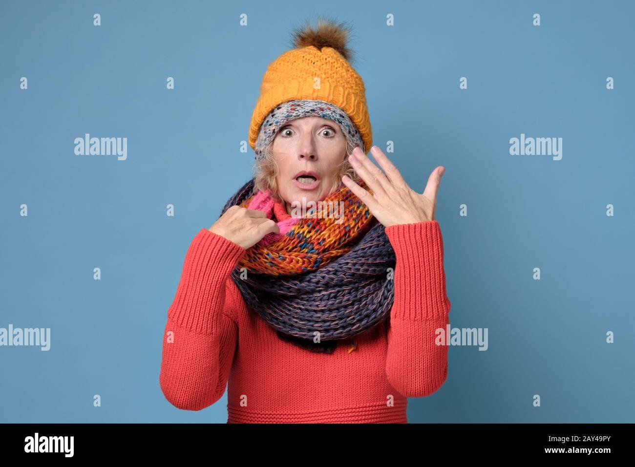 Seniorin in Hüten und Schals ist mit Schweiß bedeckt, weil sie zu viel Kleidung trägt, die an der blauen Wand isoliert ist. Kaltwetterkonzept Stockfoto
