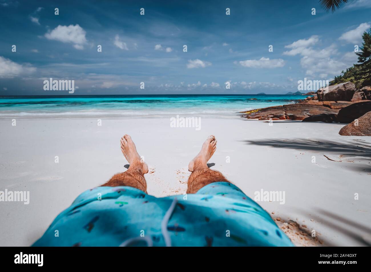 POV von Männern, die Badehosen mit gebräunten Beinen am paradiesischen weißen, tropischen exotischen Strand mit Blick auf den türkisblauen Ozean tragen. Urlaub Stockfoto
