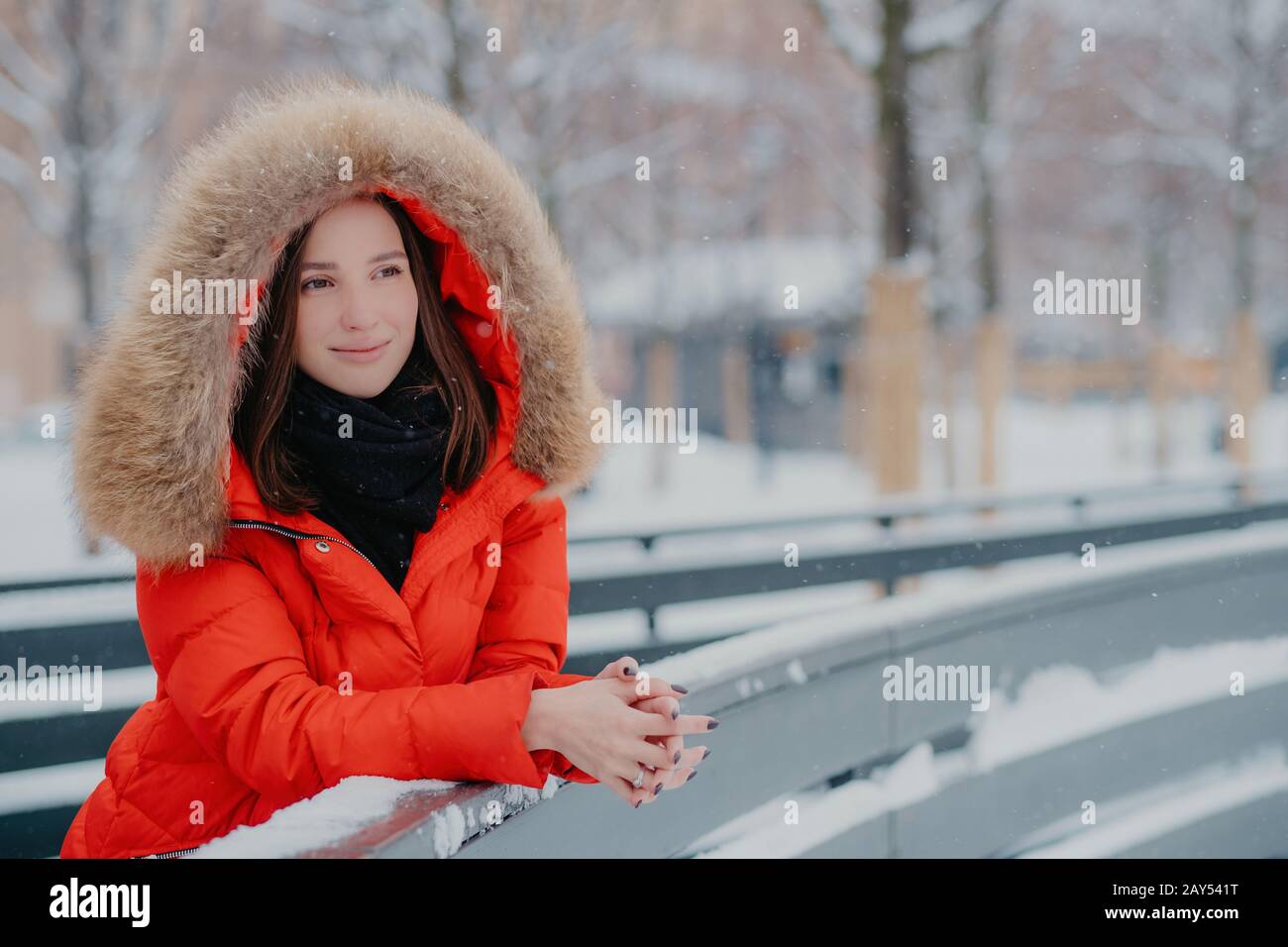 Außenaufnahme der nachdenklichen europäischen Frau trägt rote Winterjacke mit Kapuze am Kopf, lehnt sich daher an, blickt penibel in die Ferne, genießt guten Frost Stockfoto