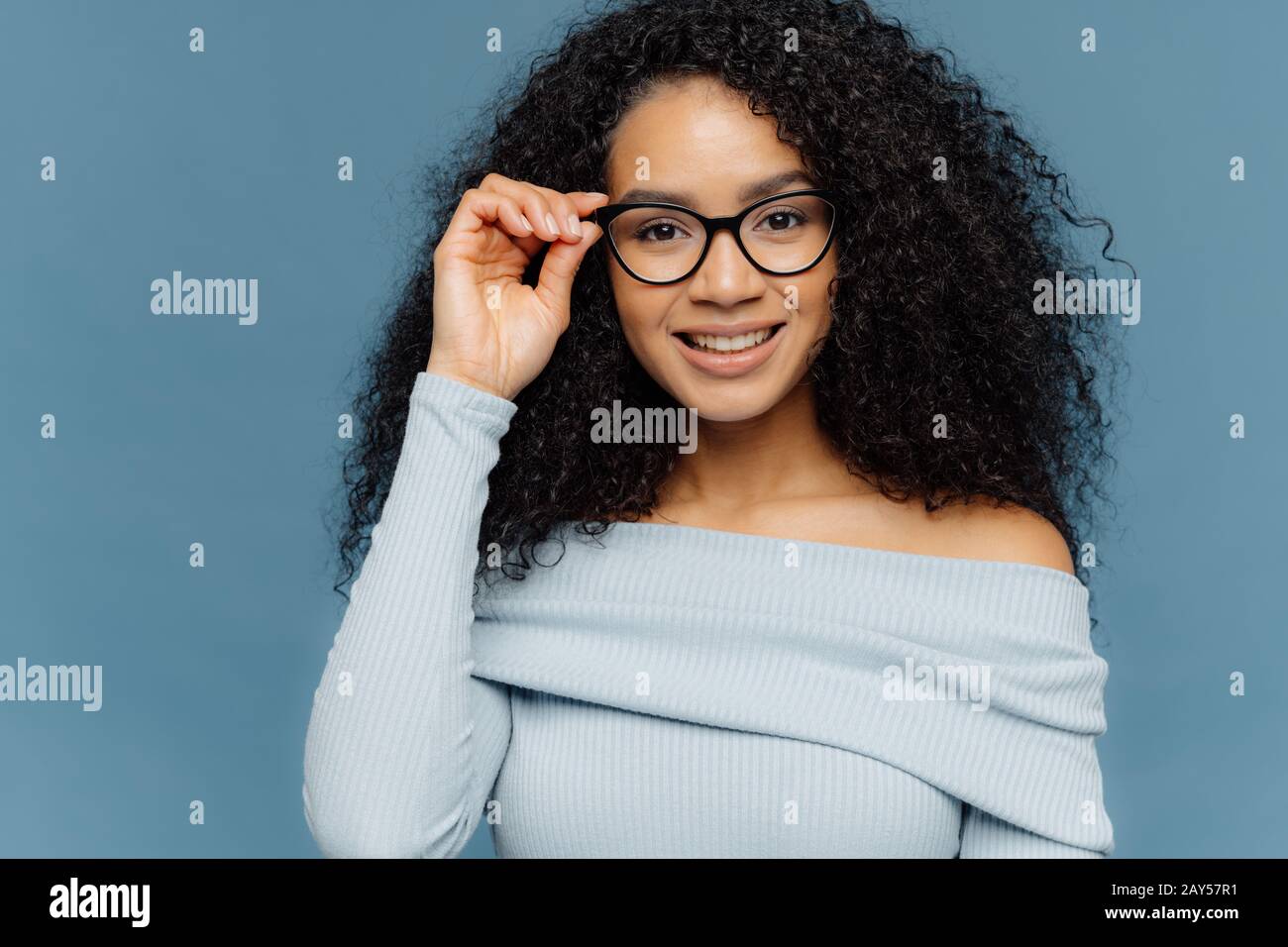 Der Kopfschuss der sympathisch aussehenden jungen afroamerikanischen Frau hält die Hand auf dem Brillenrahmen, hat ein sanftes Lächeln, trägt einen blauen Pullover, hat minimale Makeup, hea Stockfoto