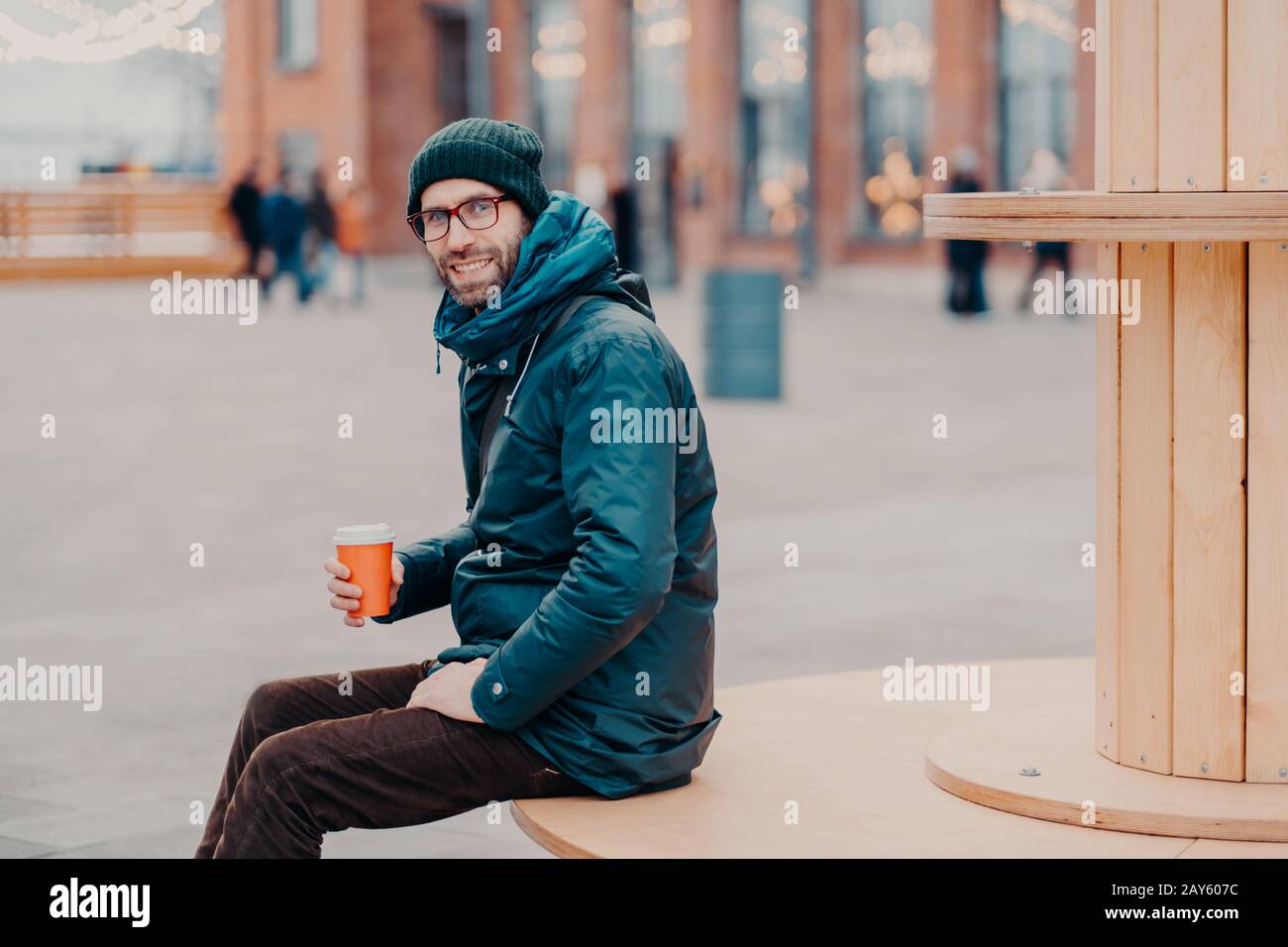 Horizontaler Blick auf den fröhlichen europäischen Mann mit dicken Borsten, trägt Hut und Jacke, hält Einwegbecher Kaffee, atmet frische Luft, posiert auf der Straße Stockfoto