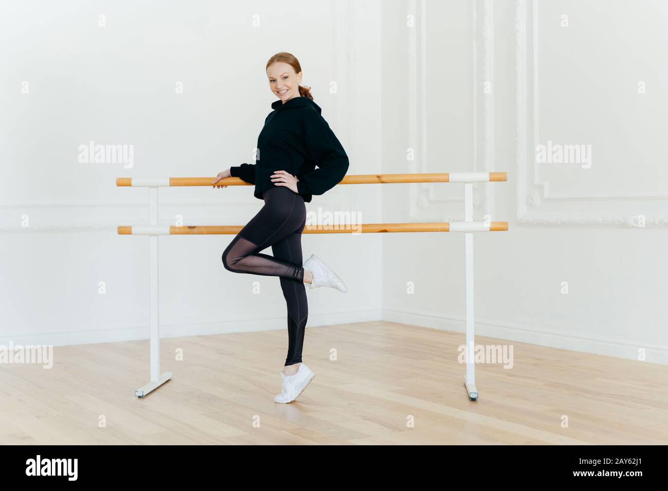 Positiv schlankes, junges Weibchen steht seitlich, posiert an einem Bein, bekleidet mit schwarzem Sweatshirt, Leggings und weißen Turnschuhen, hält Hand an Ballett barre, pe Stockfoto