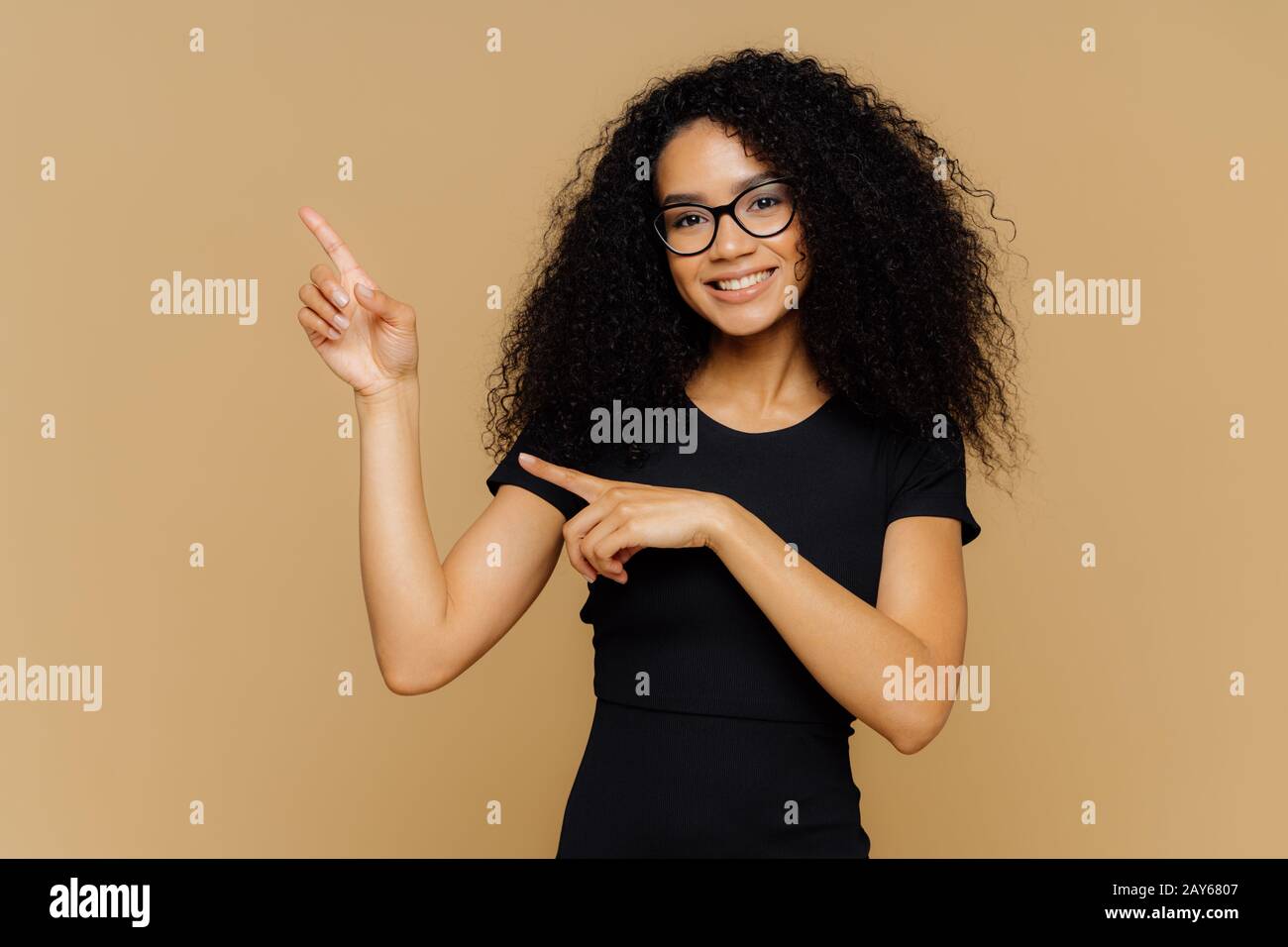 Der isolierte Schuss der glücklichen afroamerikanischen Frau zeigt sich auf und beiseite, zeigt einen leeren Kopierraum für Ihre Werbung, trägt Brille, Casual t sh Stockfoto