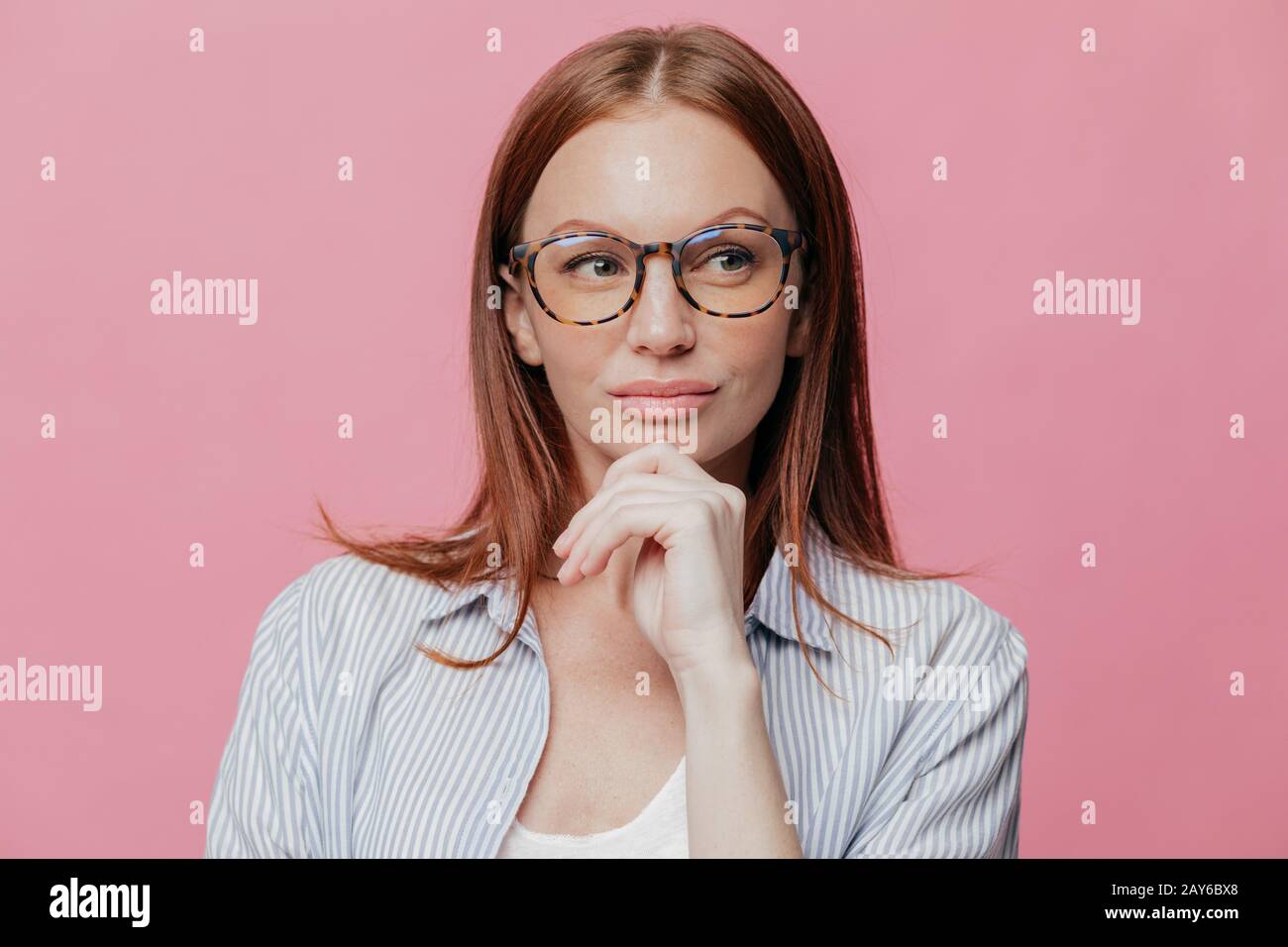 Das Foto einer nachdenklichen Frau hat einen ernsthaften Ausdruck, hält die Hand am Kinn, trägt eine Brille und ein elegantes Hemd, isoliert über dem rosafarbenen Hintergrund, denkt über Neues nach Stockfoto