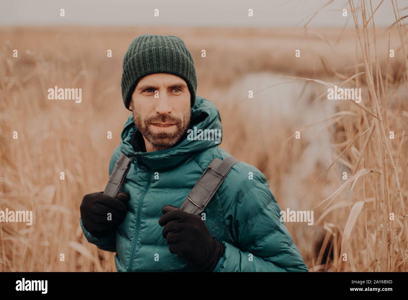 Foto eines attraktiven Mannes mit kontemplativem Ausdruck, trägt Handschuhe, Hut, wirft draußen, blickt beiseite, spaziert über Feld, denkt über etwas nach. Kalt Stockfoto
