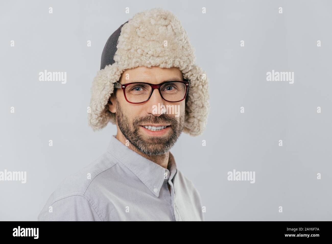 Foto von attraktiven jungen europäischen Mannes mit angenehmen Erscheinungsbild, trägt warme fut Hut, transparente Gläser, Modelle über weiße studio Wall, freut sich sho Stockfoto