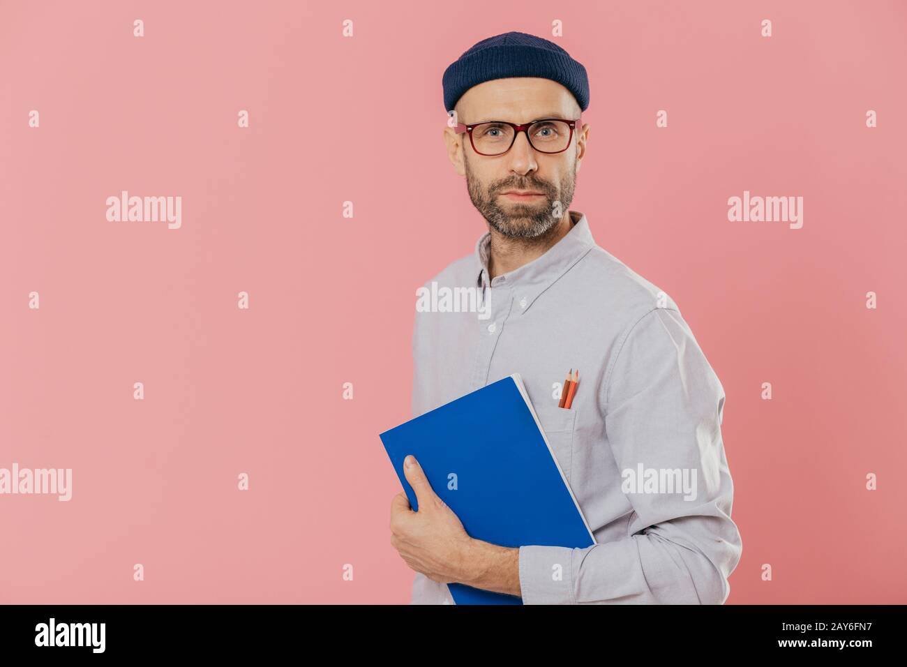 Stattliche männliche Designer trägt stilvolle Kleidung, hat zwei Stifte in der Tasche von Hemd, hält Blau notepad für Sketche und über Rosa backgroun isoliert Stockfoto
