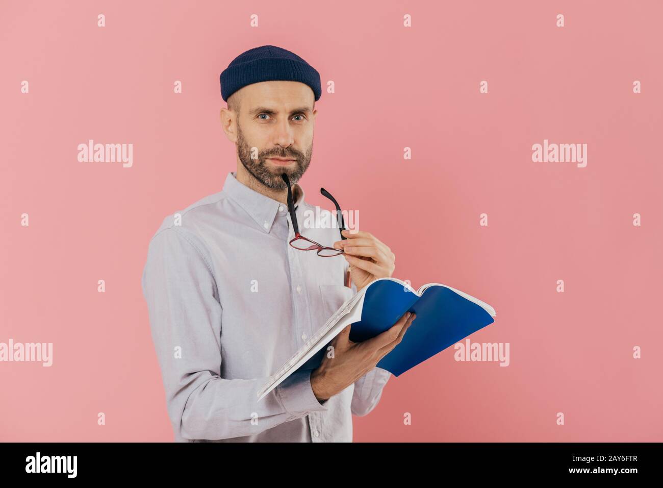 Selbstbewusst ernster Mann mit dunklen Stoppeln, nimmt Brillen, hat aufmerksamen Blick auf Kamera, hält geöffnet Lehrbuch, lernt erforderlichen Informationen, Stockfoto