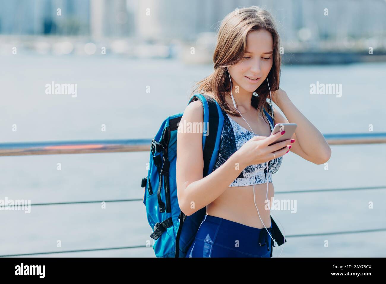 Horizontales Porträt einer hübschen Frau mit perfekter Figur, trägt Leggings und Oberteil, hat einen fokussierten Blick in das Smartphone, trägt großen Rucksack, versucht zu fi Stockfoto