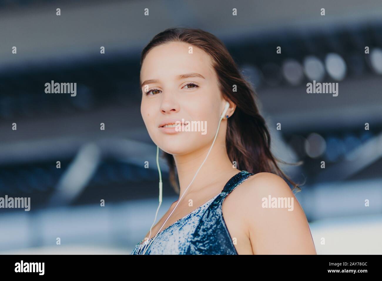 Nahaufnahme einer gut aussehenden gesunden europäischen Frau hört Musik mit unerkennbarem Gerät, blickt direkt auf die Kamera, posiert gegen verschwommenes Backgroschen Stockfoto