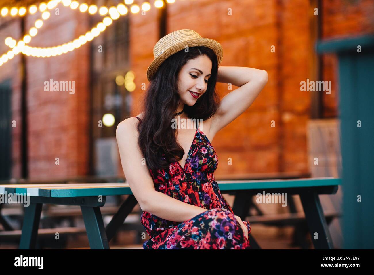Junge attraktive Frau mit europäischem Auftritt auf der Bank im Café im Freien, mit Hut und Kleid, mit der Hand hinter dem Kopf, mit Blick auf Wi Stockfoto