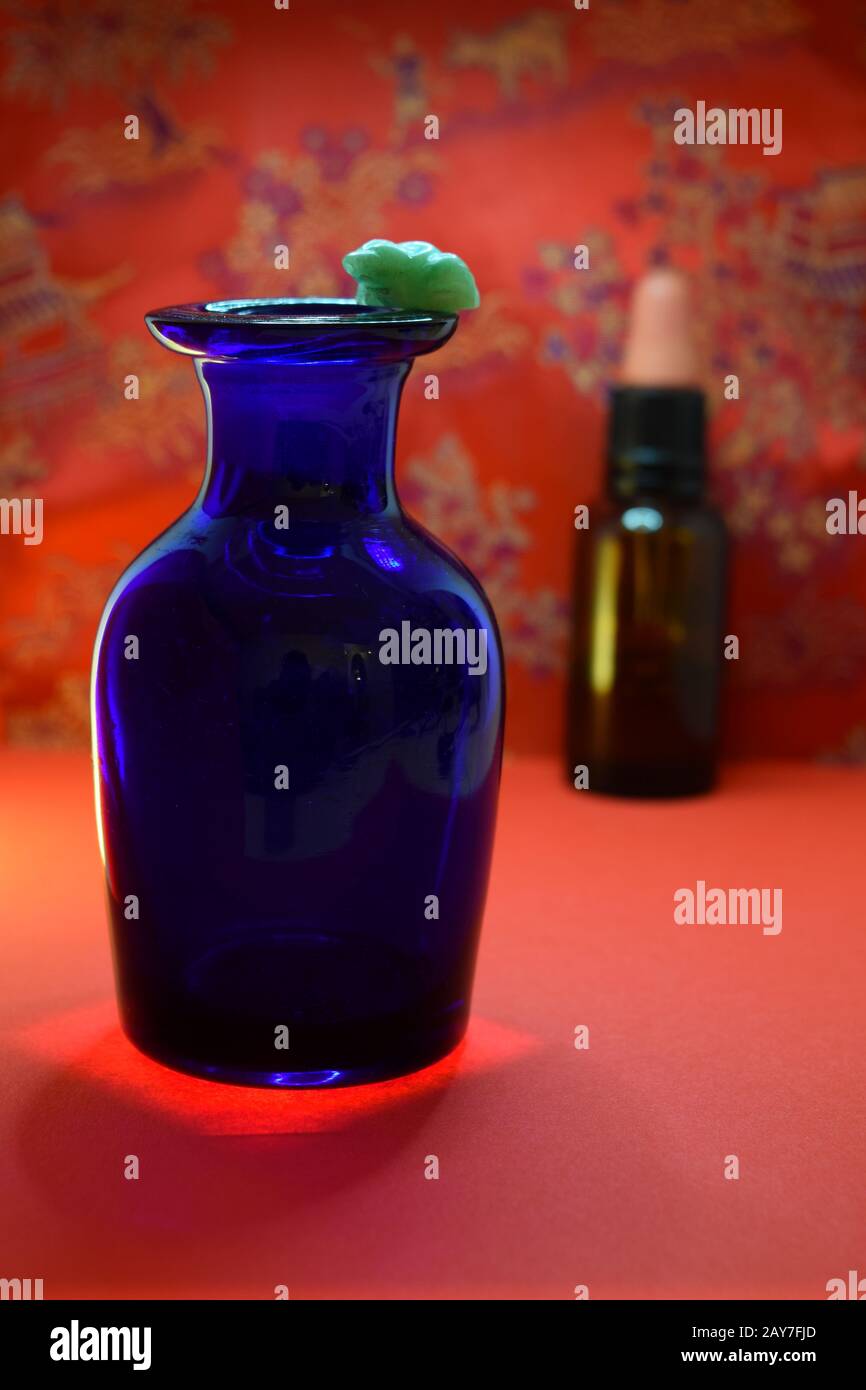 Blaue Glasflasche von unten beleuchtet mit kleinem Jade-Frosch am Rand, braune Glasflasche mit Gummistopfen im Hintergrund vor einem rot-chinesischen Muster. Stockfoto