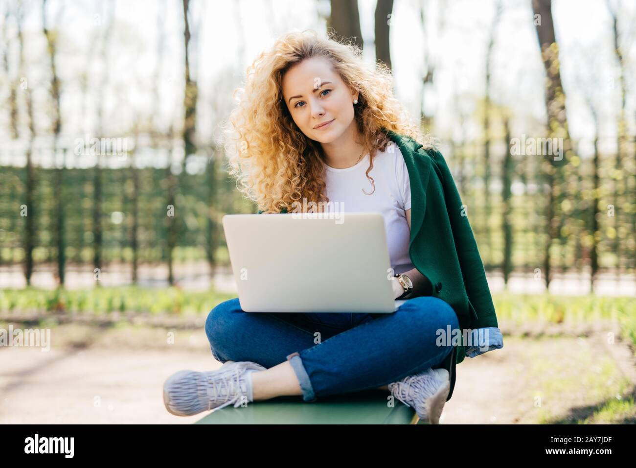 Kaukasische Frau mit lockig hellem Haar und blauen Augen, die bequeme Kleidung trägt, die gekreuzte Beine im Freien sitzt und den Laptop auf den Knien hält, Nachrichtenübermittlung w Stockfoto