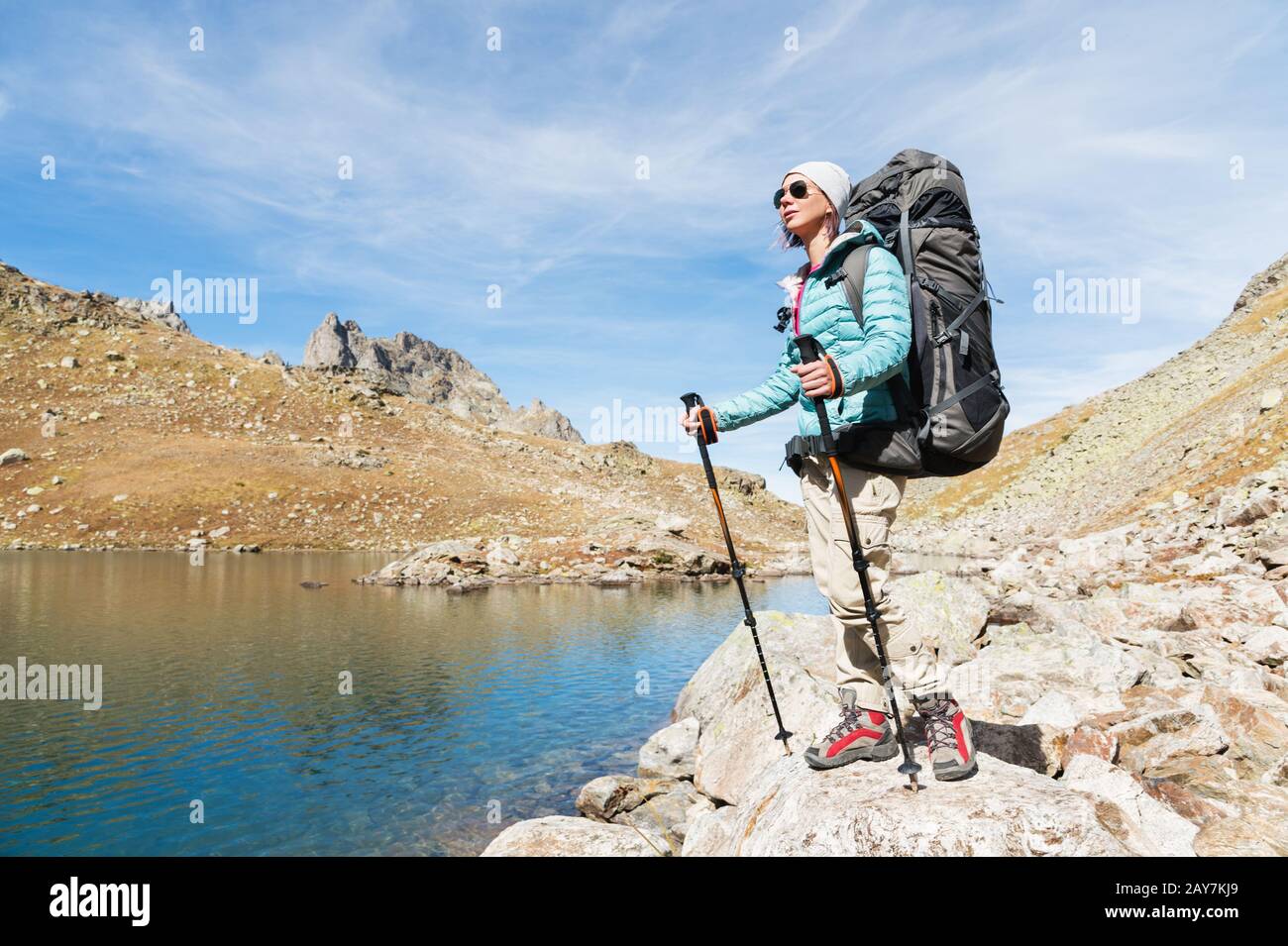 Ein Wandermädchen in Sonnenbrille und ein Hut mit Rucksack und Bergausrüstung mit aufspürbaren Trecks in den Händen blickt auf das schöne V Stockfoto