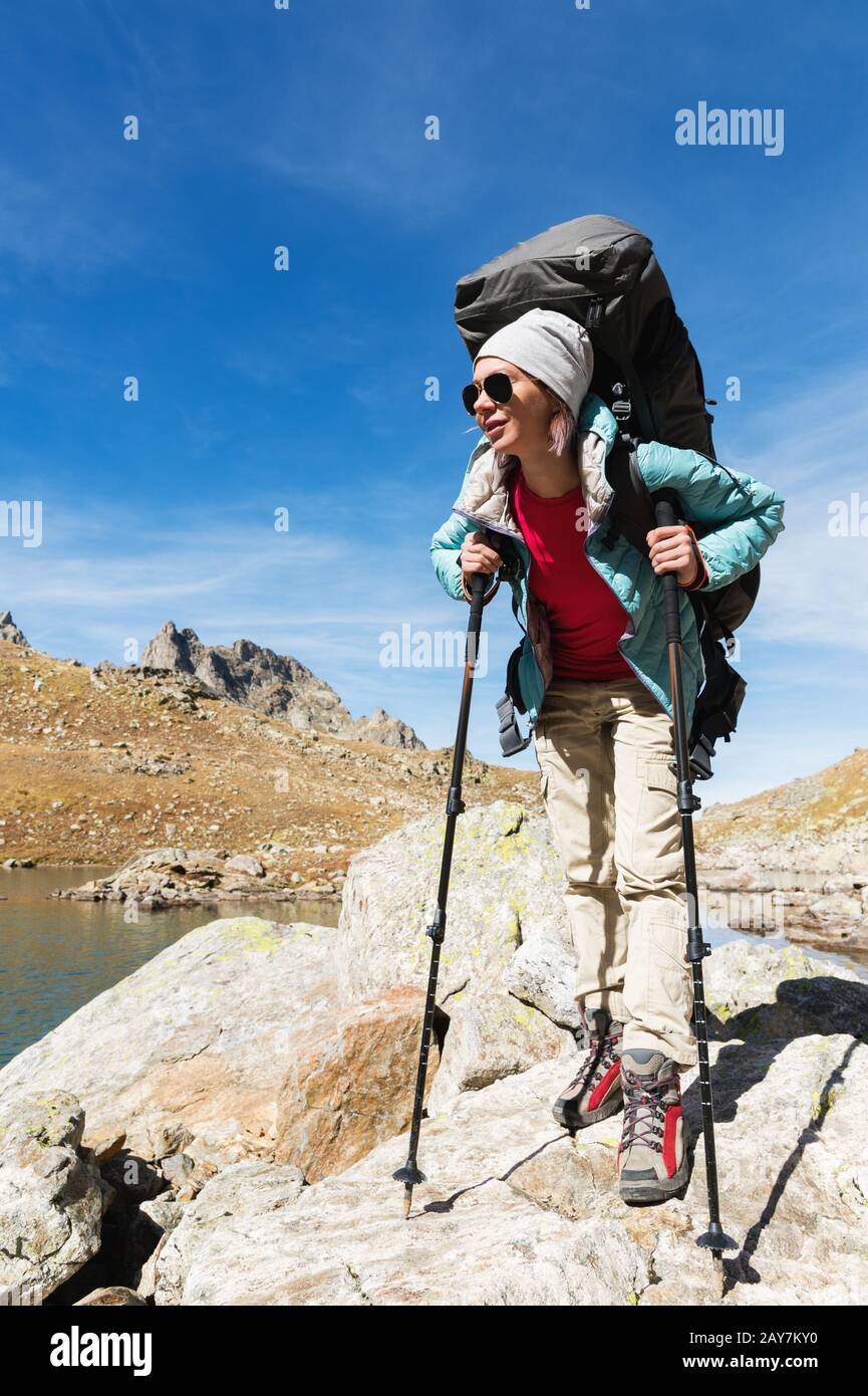 Ein Wandermädchen in Sonnenbrille mit Rucksack und Tracking-Stöcken steigt vor dem Hintergrund von Felsen und einer Höhe von auf einen hohen Felsen Stockfoto