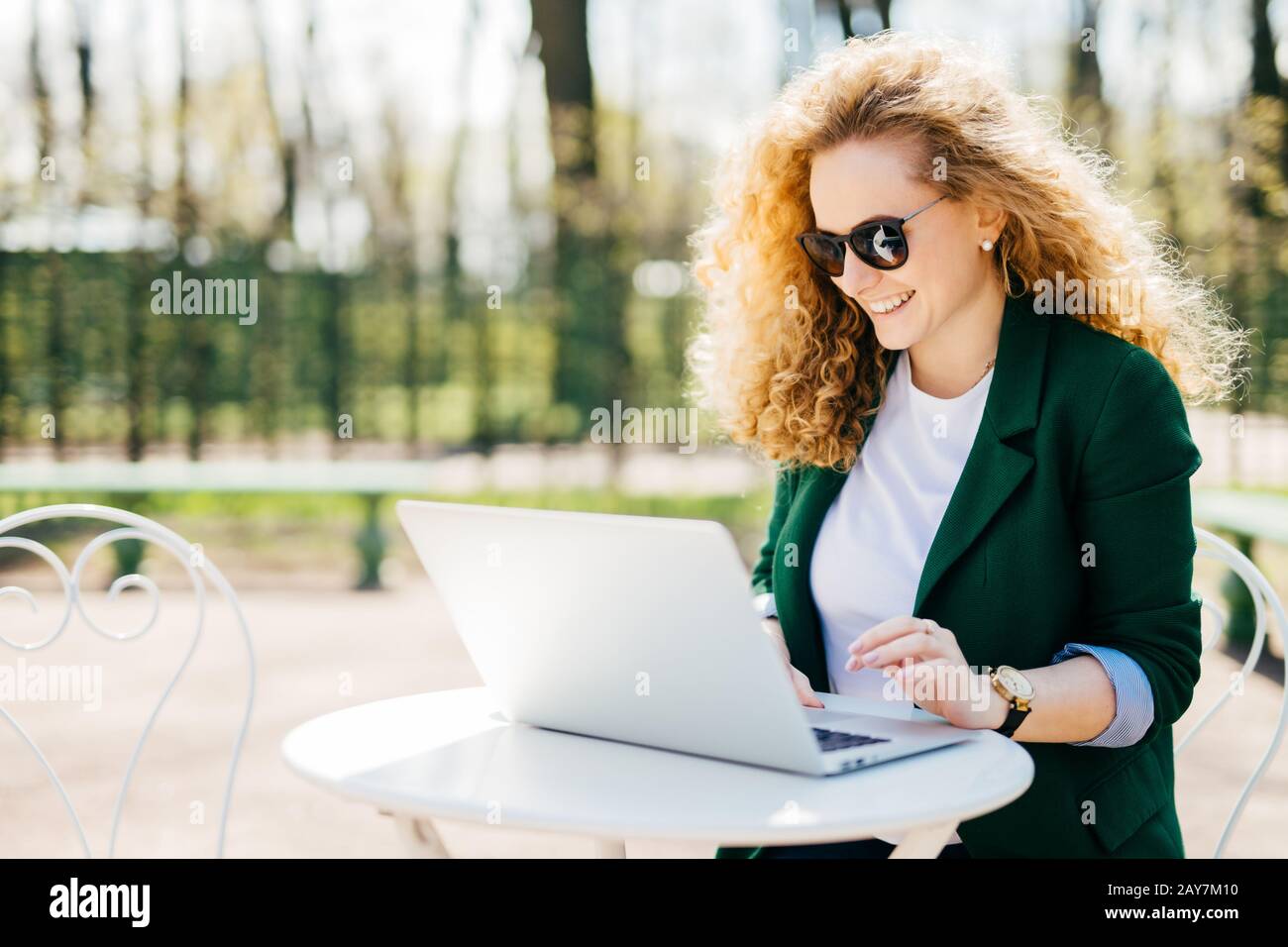 Außenporträt von attraktiven jungen, lockigen Frauen, die am Schreibtisch im Park sitzen, indem sie ihren Laptop verwenden, um Nachrichten an ihren Freund zu schreiben, der gerne Somet liest Stockfoto