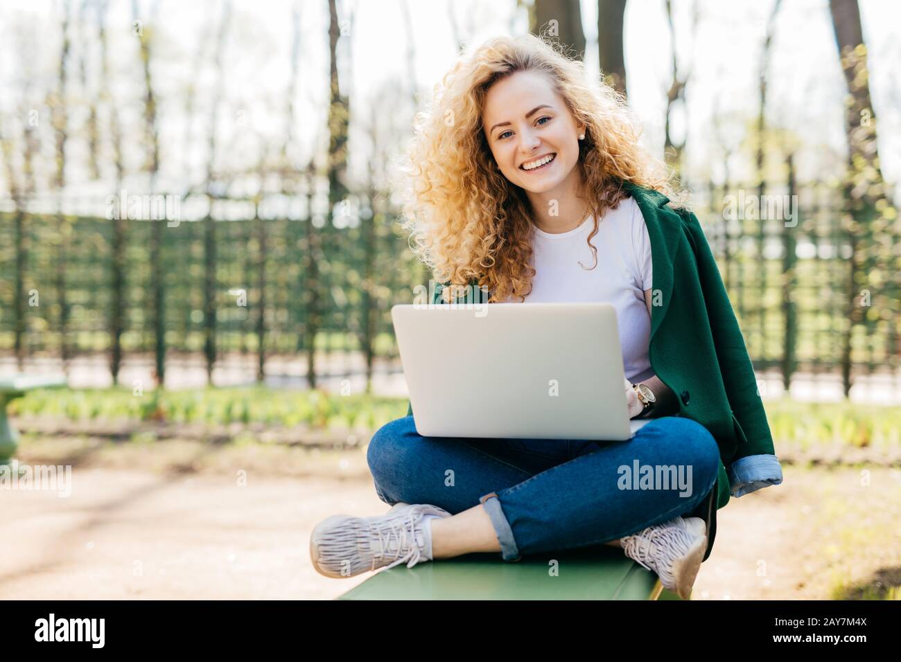 Gefiel der europäischen Frau, die schöne, flauschige blonde Haare hat, die Jeans, Jacke und Sportschuhe trägt, die gekreuzte Beine mit einem Laptop-Computer für Kommun sitzen Stockfoto