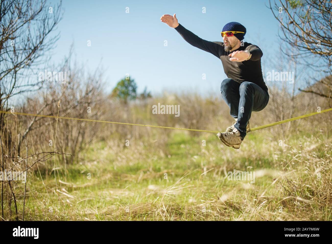 Ein Mann im Alter, der mit einem Hut und Turnschuhen auf der Slackline in einer Brille sitzt, fängt das Gleichgewicht ein und genießt das Leben auf der Natur i Stockfoto