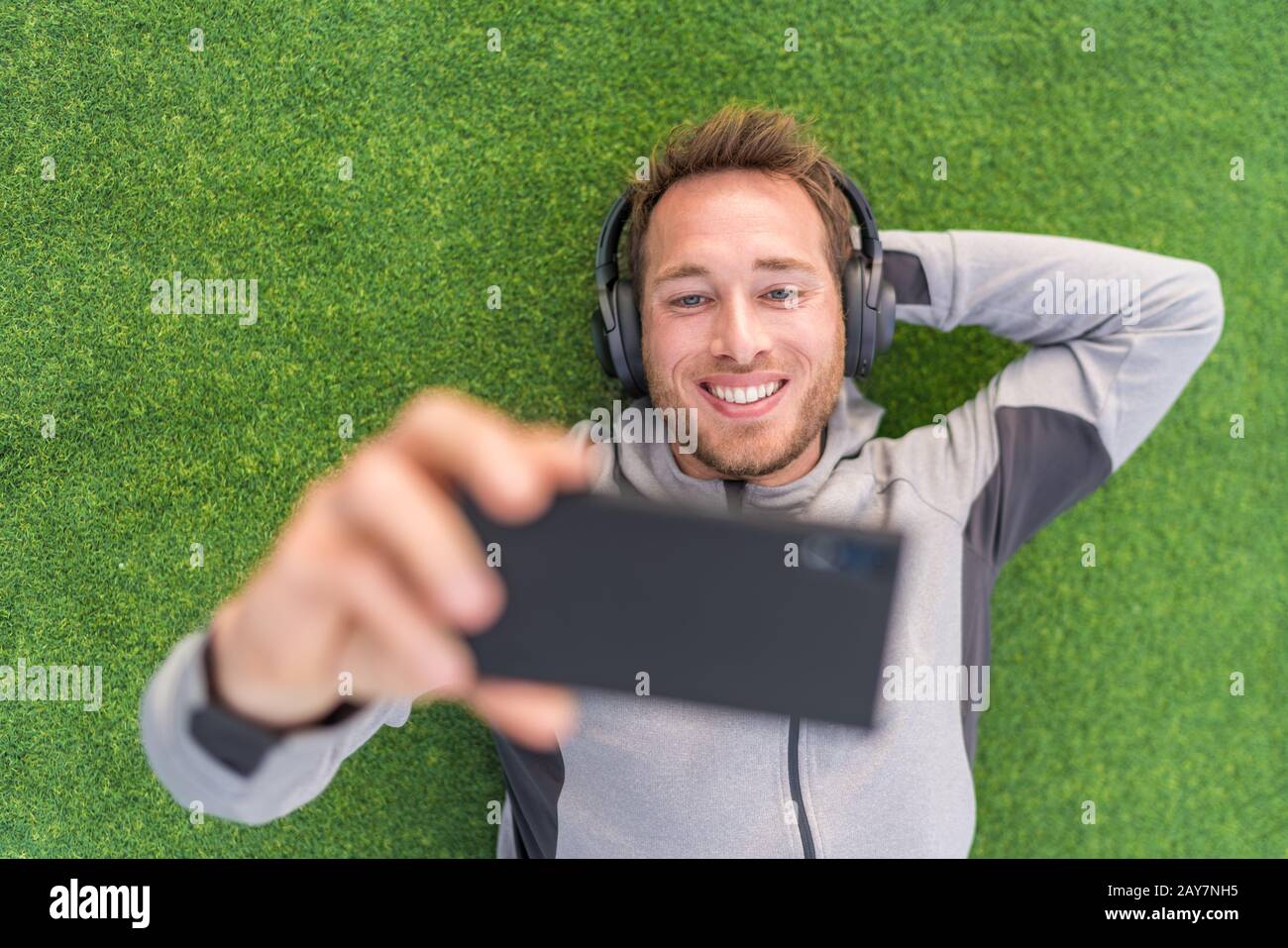 Happy man sieht Telefon-Video-App mit Kopfhörern und Smartphone draußen auf dem grünen Sommergras entspannen. Stockfoto