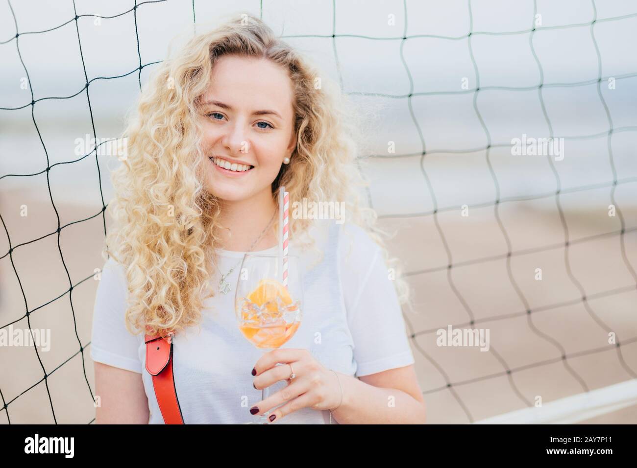 Die entzückende junge Europäische Frau hat buschige, lockige Haare, lächelt sanft, hält ein Glas Orangencocktail, steht in der Nähe des Tennisnetzes mit Kopierraum rechts f Stockfoto