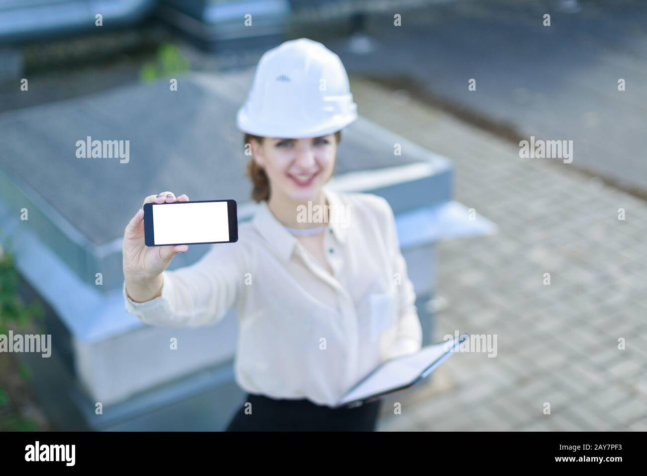 Attraktive Geschäftsfrau in weißer Bluse, Armbanduhr, Helm und schwarzem Rock stehen auf dem Dach und zeigen leeres Smartphone Stockfoto