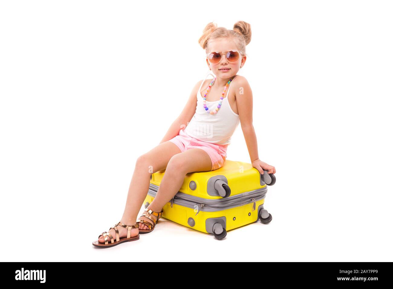 Auf dem gelben Koffer sitzen süßes junges Mädchen in weißem Hemd, rosafarbene Shorts und Sonnenbrille Stockfoto